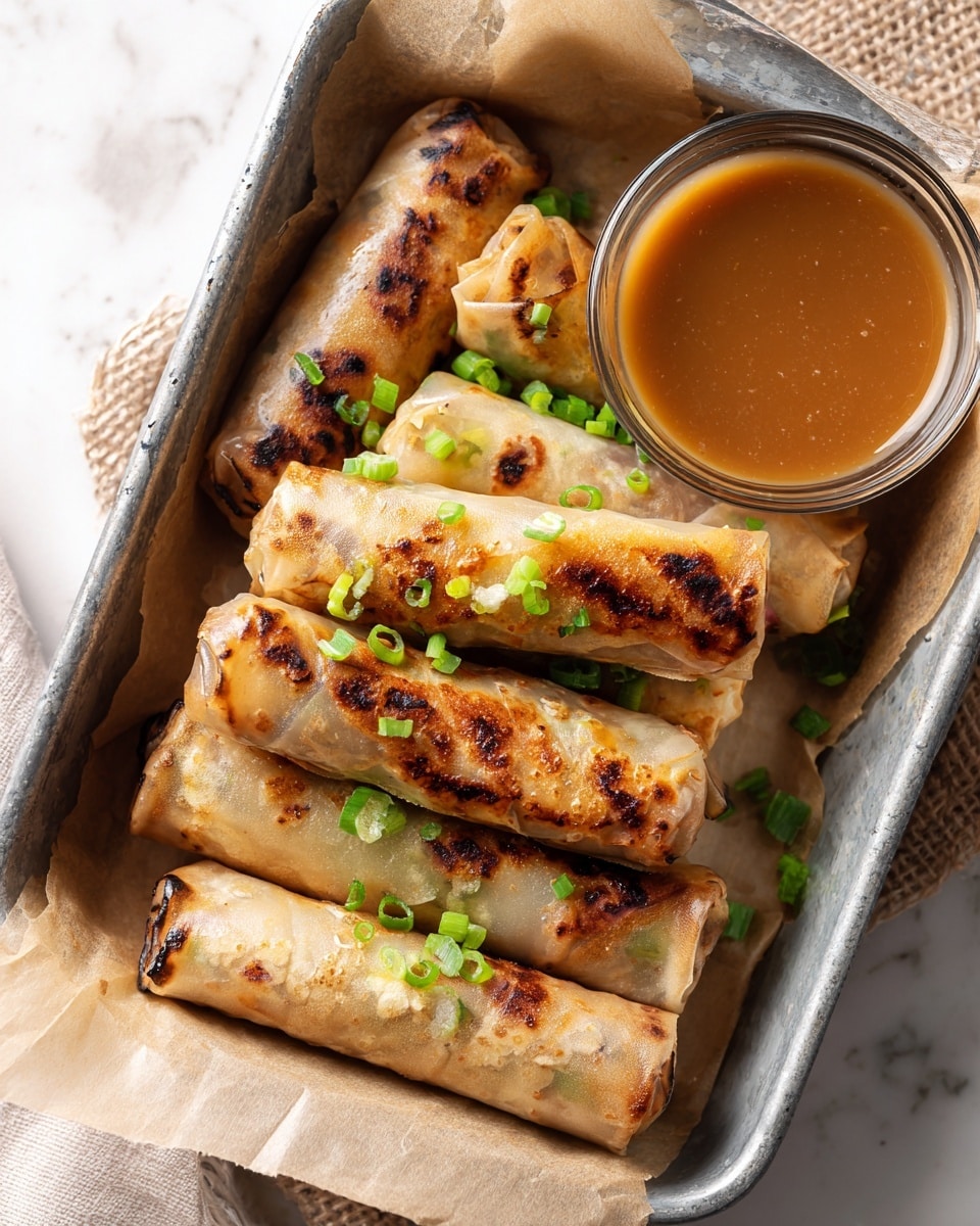 The image shows six translucent spring rolls with a light golden-brown color and some charred spots, arranged close together in a metal tray lined with parchment paper on a white marbled surface. Each roll has a slightly wrinkled texture and is garnished with small pieces of bright green chopped scallion on top. A clear glass bowl of smooth, medium-dark brown dipping sauce sits on the right side of the tray, partially visible. The scene is warm and inviting with a focus on the textures of the spring rolls and sauce. Photo taken with an iphone --ar 4:5 --v 7