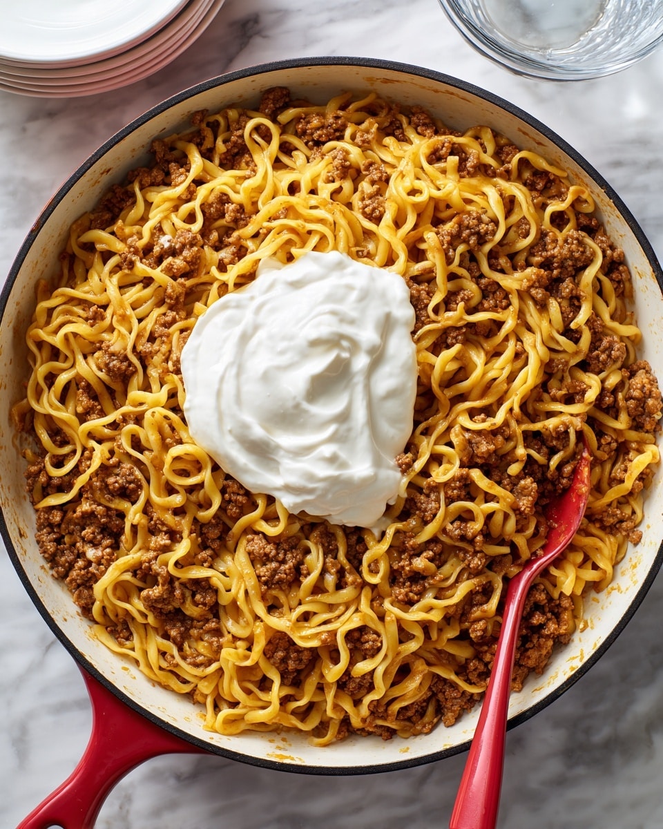 A large white pan with a black rim holds a cooked dish of yellow egg noodles mixed with browned ground meat, evenly spread across the pan. On top in the center is a dollop of thick white sour cream. A red spoon with some sour cream and sauce on it rests inside the pan on the right side. The pan is placed on a white marbled surface with a small part of a clear glass bowl and a white bowl partially visible in the background. Photo taken with an iphone --ar 4:5 --v 7