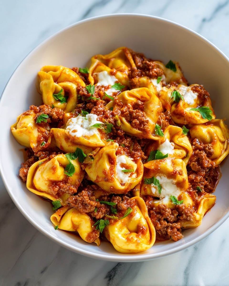 A close-up image of a white bowl filled with tortellini pasta in a thick, red meat sauce. The tortellini are yellow with a slightly glossy texture, folded into ring shapes and partly covered with browned ground meat. There are small melted white cheese patches spread unevenly on top. Small green parsley leaves are scattered across the dish, adding color. The bowl sits on a white marbled surface, and natural light softly highlights the textures and colors of the food. Photo taken with an iphone --ar 4:5 --v 7