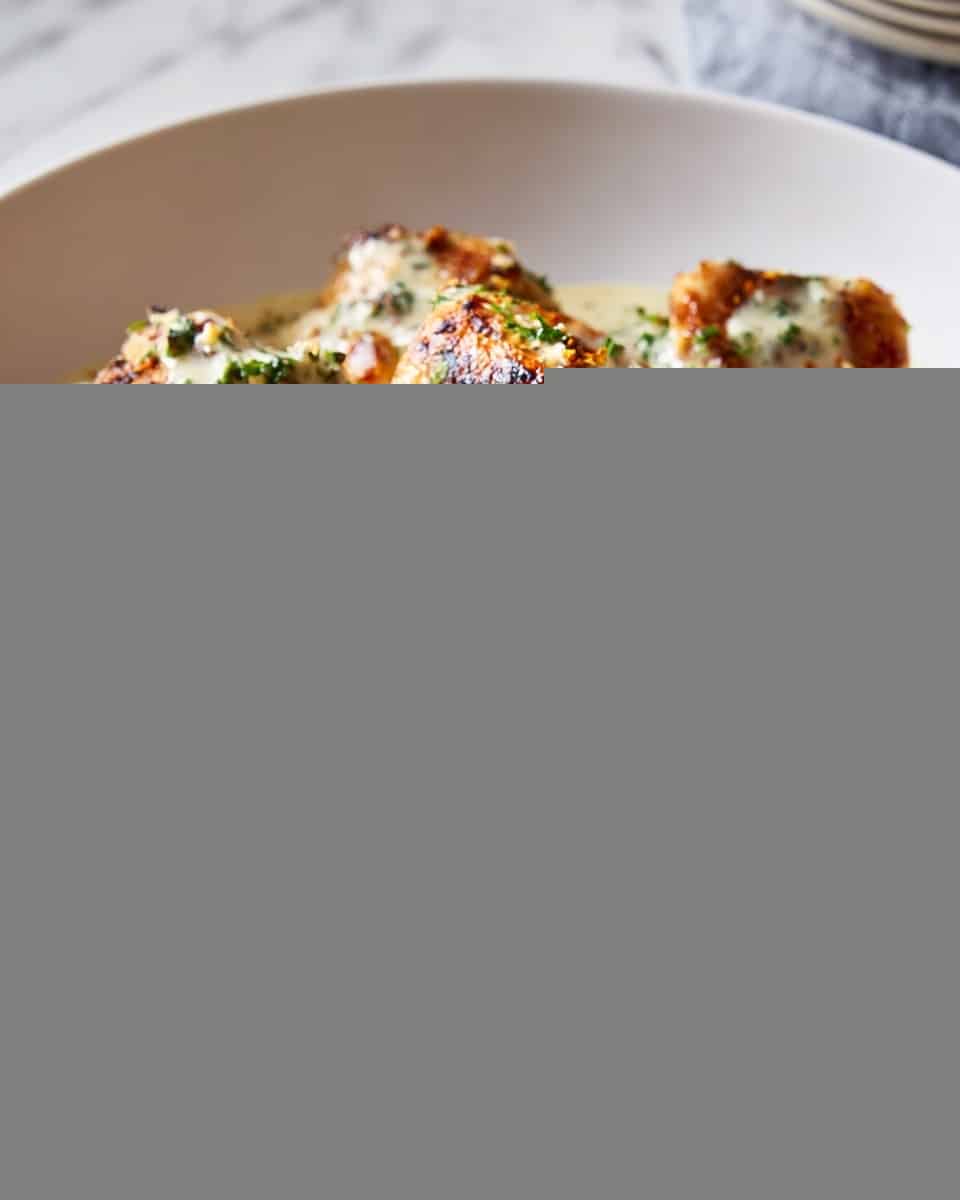A white bowl is filled with many pieces of grilled chicken, golden brown with darker charred spots, all covered in a creamy, pale sauce flecked with green herbs. The chicken pieces sit on top, with the sauce pooling beneath and around them, creating a rich, textured look. The bowl is placed on a white marbled surface with more dishes slightly blurred in the background. photo taken with an iphone --ar 4:5 --v 7