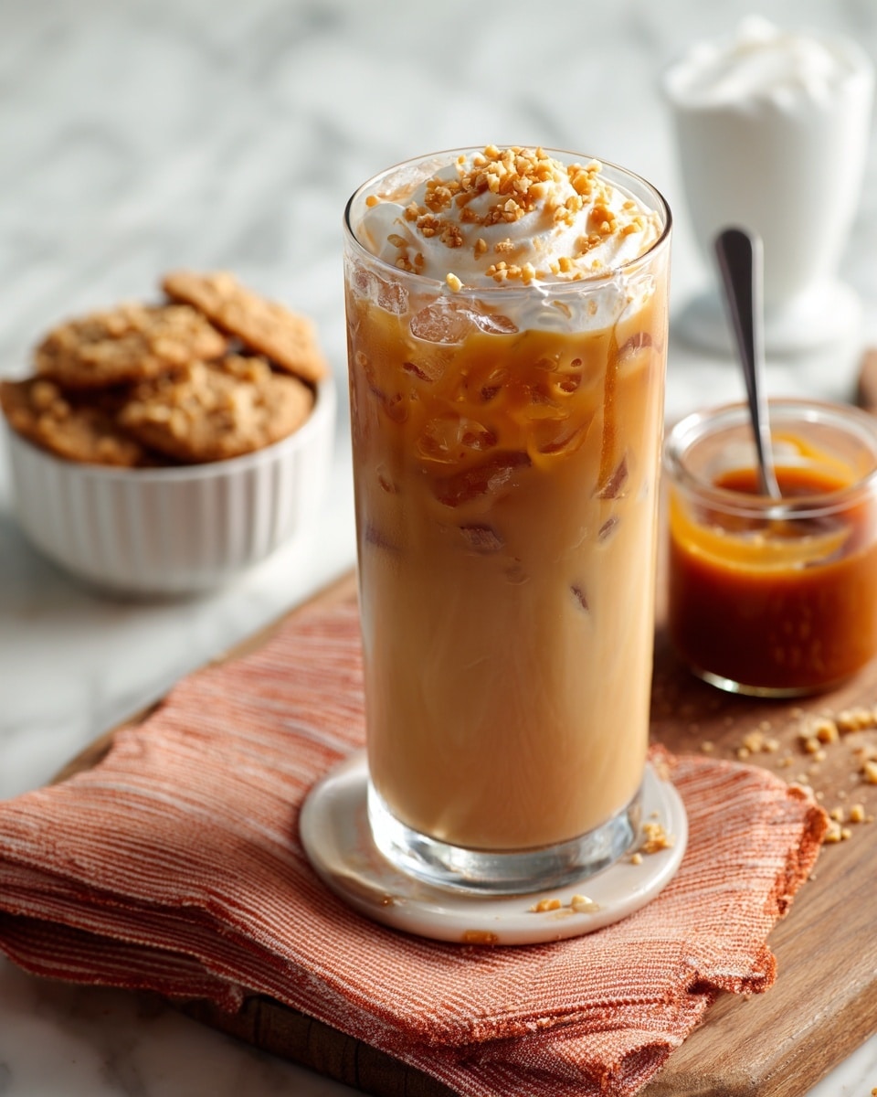 The image shows a tall clear glass filled with iced coffee mixed with cream, giving it a light beige color with visible ice cubes inside. The drink is topped with whipped cream and sprinkled with crushed nuts. This glass is placed on a wooden board alongside a small white bowl filled with cookies on an orange and white striped cloth. A small glass jar containing caramel sauce is nearby with a spoon inside. The background and surface are a white marbled texture. photo taken with an iphone --ar 4:5 --v 7