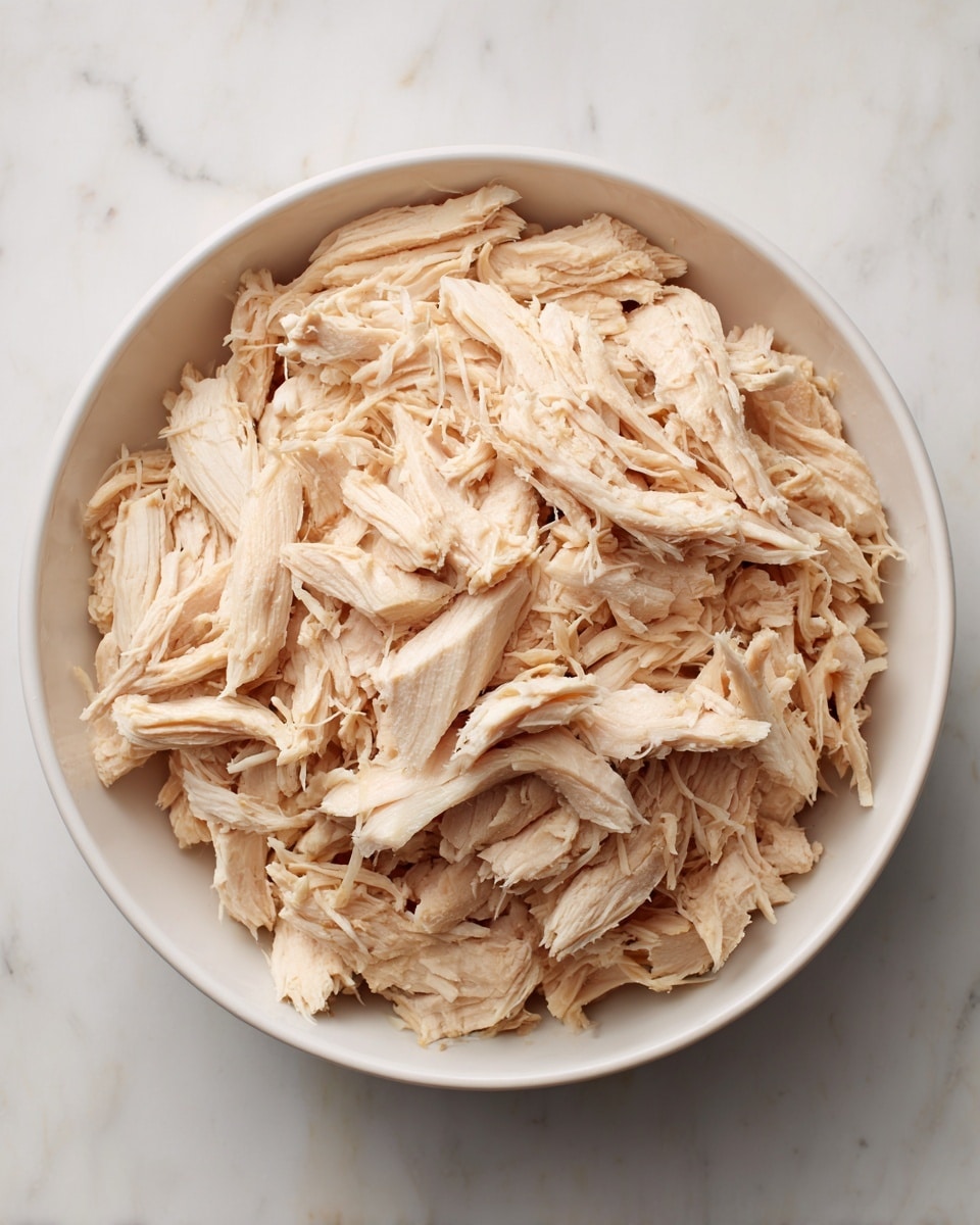 A white bowl filled with shredded cooked chicken that looks soft and tender, with light brown and beige tones showing the fibers of the meat. The chicken pieces are spread evenly inside the bowl, with some small bits and strings of meat scattered on top. The bowl sits on a white marbled surface. photo taken with an iphone --ar 4:5 --v 7