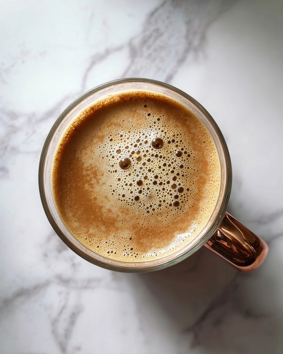 A clear glass cup with a bronze handle is filled with a light brown foamy liquid, showing a smooth, frothy layer with small bubbles on the top. The cup is placed on a white marbled surface with gray veins, and the image is taken from above, focusing on the creamy texture inside the cup. photo taken with an iphone --ar 4:5 --v 7