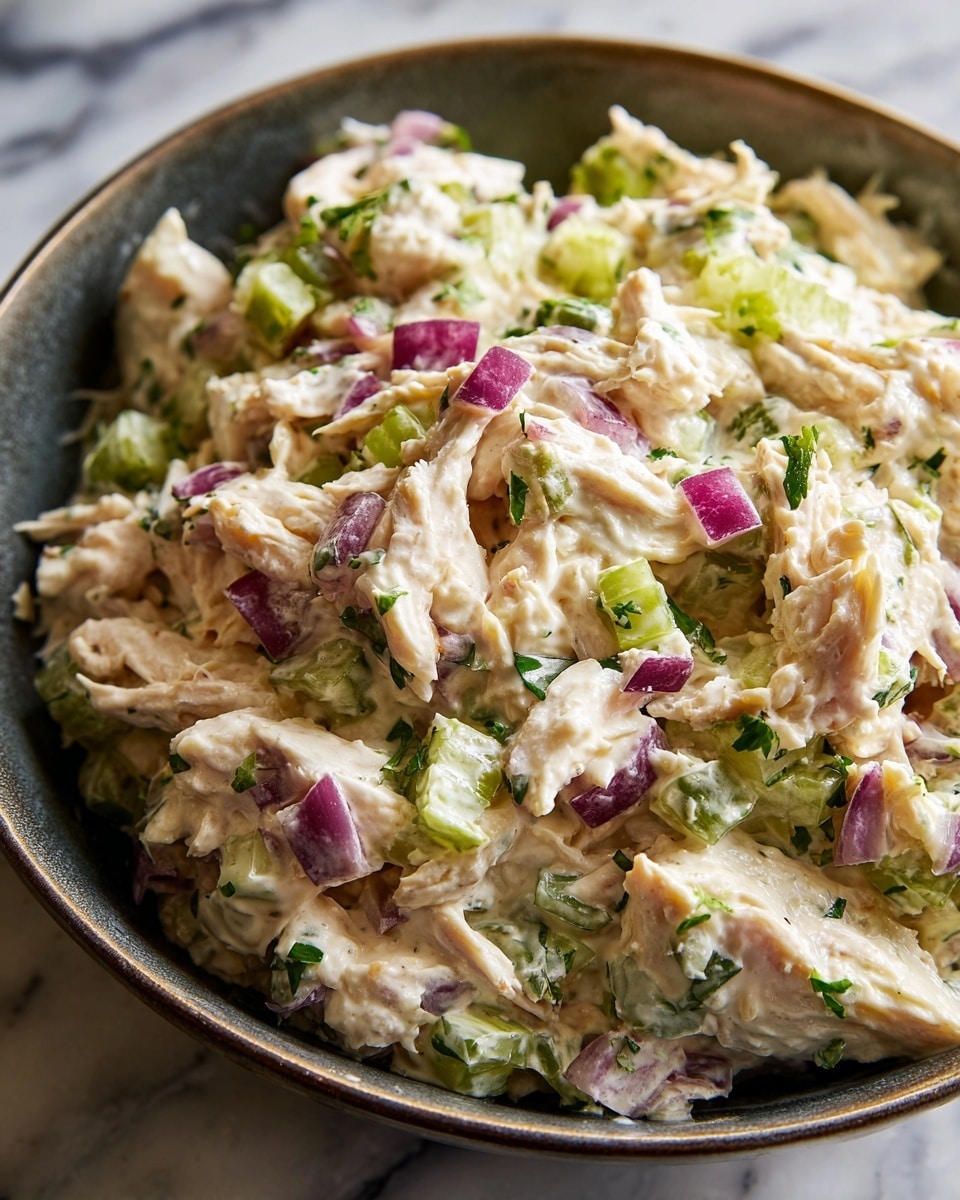 A close-up view of a creamy salad served in a dark bowl placed on a white marbled surface, showing a mixture with three main layers: the first layer is shredded light beige chicken mixed with white creamy dressing, the second layer contains small pieces of red onion with purple skin and white inside, and the third layer shows chopped green vegetables like celery and herbs, all combined to create a moist and textured appearance with small leafy bits scattered throughout, filling the bowl almost to the top; photo taken with an iphone --ar 4:5 --v 7