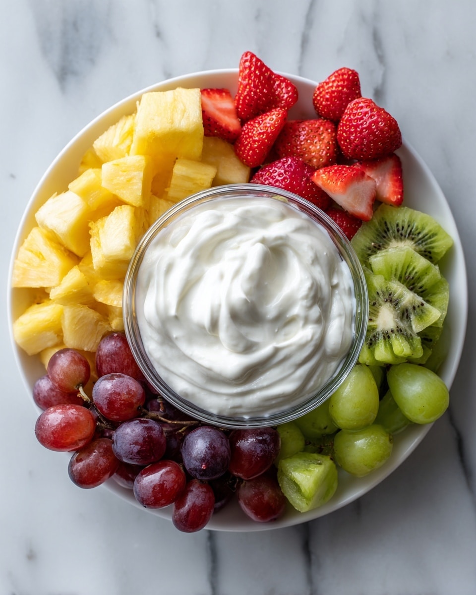 A white plate holds a small clear glass bowl filled with a thick, creamy white yogurt that has a smooth, swirled texture on top. Around the bowl is a colorful mix of fresh fruits arranged in layers: bright yellow pineapple cubes, red grapes, green grapes, sliced strawberries showing their juicy red inside, and slices of green kiwi with their dark seeds visible. The plate sits on a white marbled surface. photo taken with an iphone --ar 4:5 --v 7