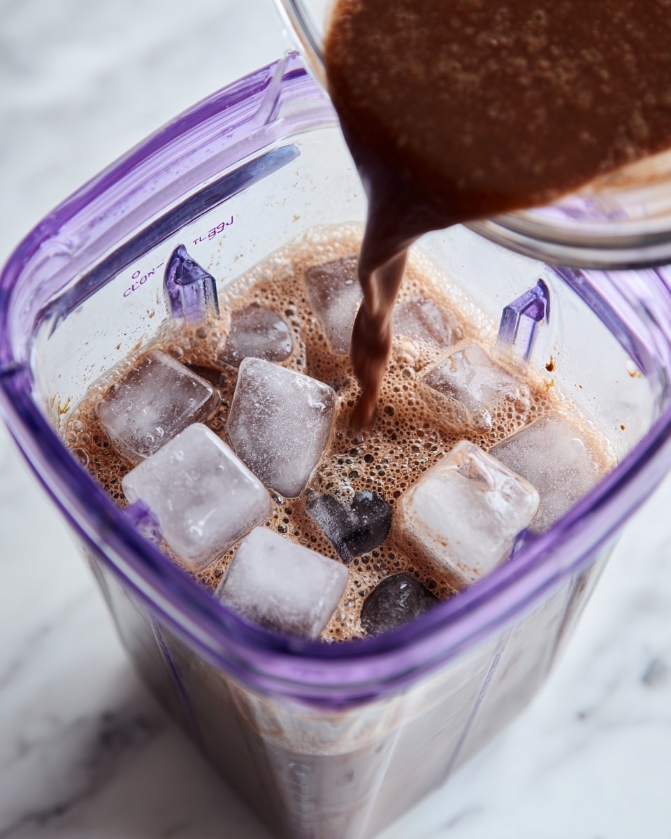 A clear blender container filled halfway with large ice cubes that are light gray and translucent, and a thick chocolate liquid being poured into it from above; the chocolate layer is dark brown and frothy with bubbles, mixing with the ice inside the clear container that has a purple tint on its edges, all set against a white marbled surface. photo taken with an iphone --ar 4:5 --v 7