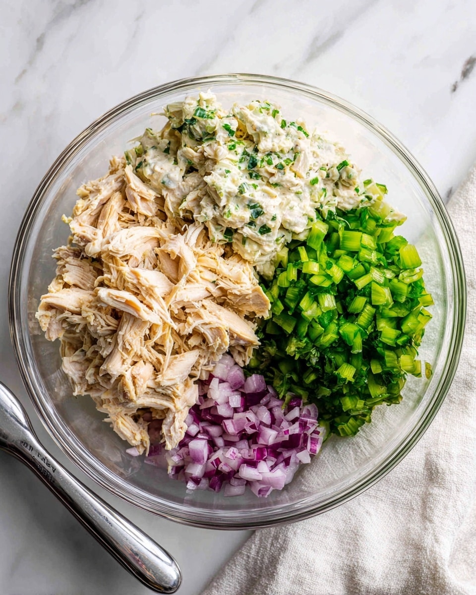 The image shows a clear glass bowl with four layers placed side by side inside: shredded cooked chicken in light beige color with black specks at the bottom left, creamy white sauce with green herb bits on top of the chicken on the left, bright green chopped celery on the top right, and small chopped purple onion pieces on the bottom right. The bowl sits on a white marbled surface with a white and gray cloth partially tucked under it on the right side. Part of a woman's hand holding a silver tongs is visible near the bottom left edge of the bowl. Photo taken with an iphone --ar 4:5 --v 7