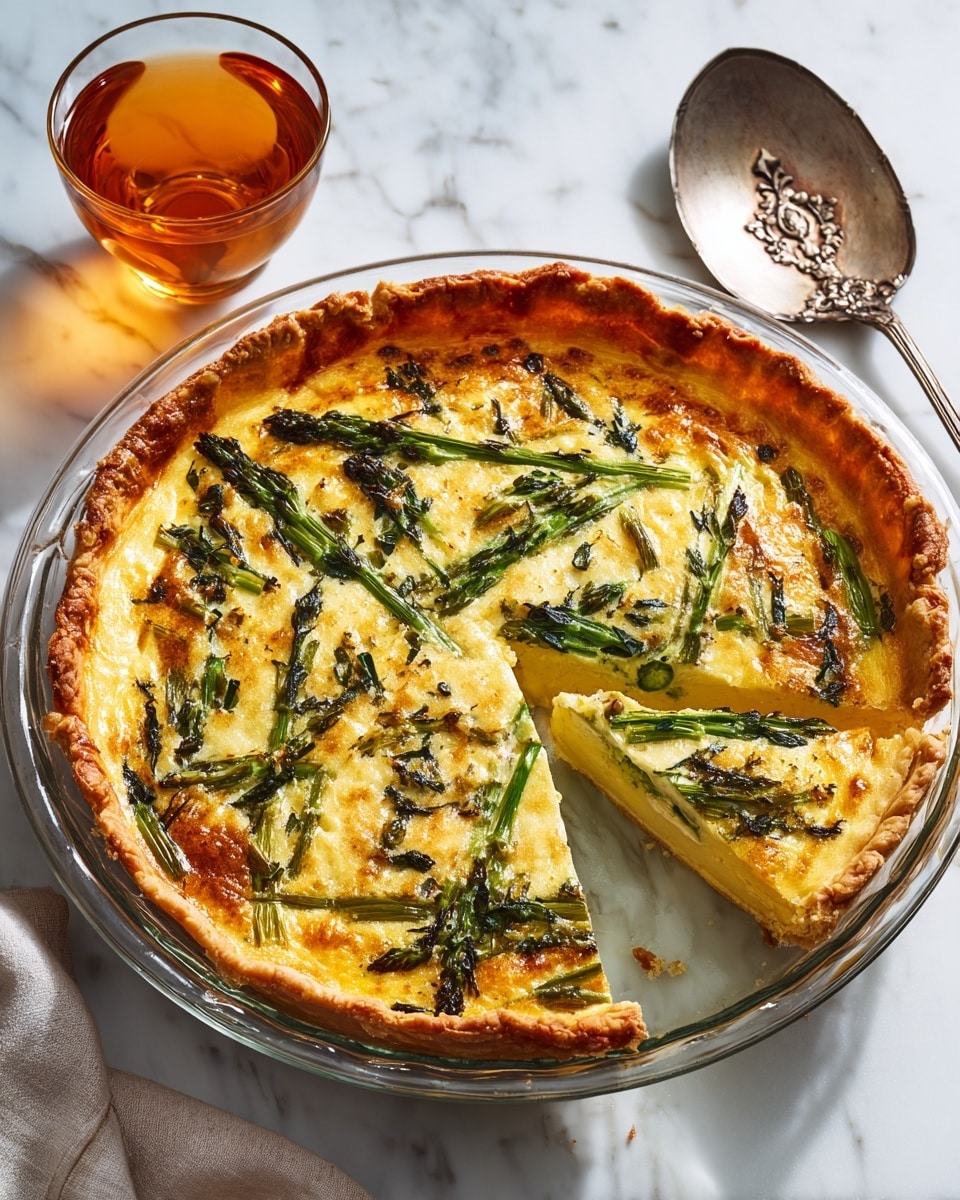 The image shows a round quiche with a golden crust on the edges, placed inside a clear glass pie dish. The quiche has two clear layers: the bottom layer is a light brown crust, and the top layer is filled with a creamy yellow egg mixture mixed with scattered pieces of dark green spinach leaves and light green asparagus pieces. There is a slice missing from the quiche, revealing the smooth texture inside. To the right of the dish is a large silver knife with a detailed handle on a white marbled surface, and above the dish is a glass cup with amber-colored liquid. The photo was taken with an iphone --ar 4:5 --v 7
