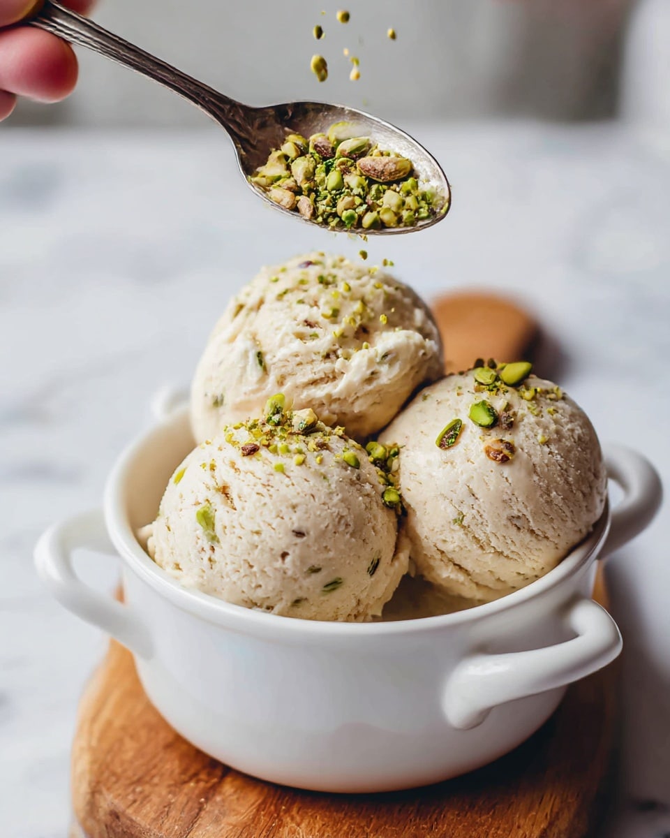 A white bowl with two small handles holds three scoops of creamy light beige ice cream with small brown bits visible inside. Each scoop is topped with a sprinkle of chopped bright green and brown pistachios, adding a crunchy texture. A silver spoon held by a woman's hand is placing more chopped pistachios on the top scoop. The bowl is set on a round wooden board, while the background is a soft white marbled surface with blurred pistachio nuts and a glass bottle in the back. photo taken with an iphone --ar 4:5 --v 7