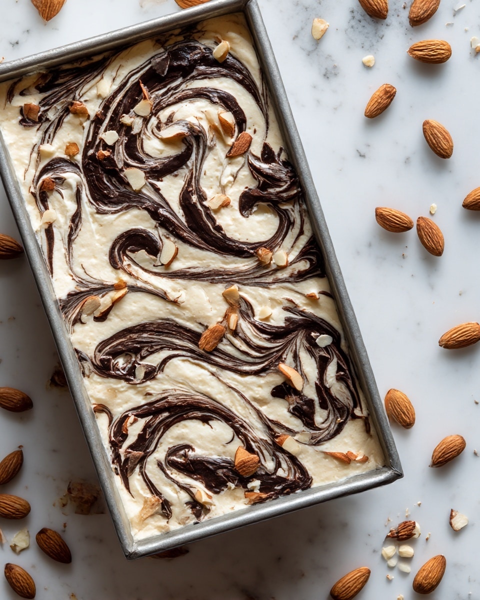 A close-up of a dessert in a silver metal rectangular pan with a creamy beige top layer swirled with dark chocolate in curly patterns, scattered with whole and chopped almonds. The pan sits on a white marbled surface with some whole almonds nearby. The texture of the top layer is smooth and slightly shiny, showing clear chocolate swirls. Photo taken with an iphone --ar 4:5 --v 7