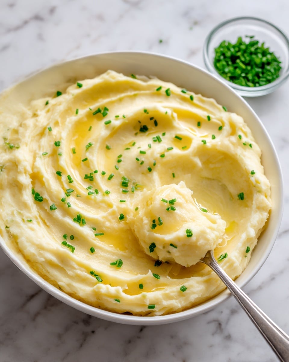 A white plate holds a generous mound of creamy mashed potatoes with a smooth and fluffy texture, topped with small green chive pieces scattered on top and around it. In the center, there is a small pool of melted butter adding a glossy yellow touch. To the right, a silver spoon rests partially in the mashed potatoes. In the background, a white casserole dish filled with more mashed potatoes is visible, with a silver spoon resting inside. Next to it, there are two small clear bowls containing chopped herbs. The whole scene is set on a white marbled surface. Photo taken with an iphone --ar 4:5 --v 7