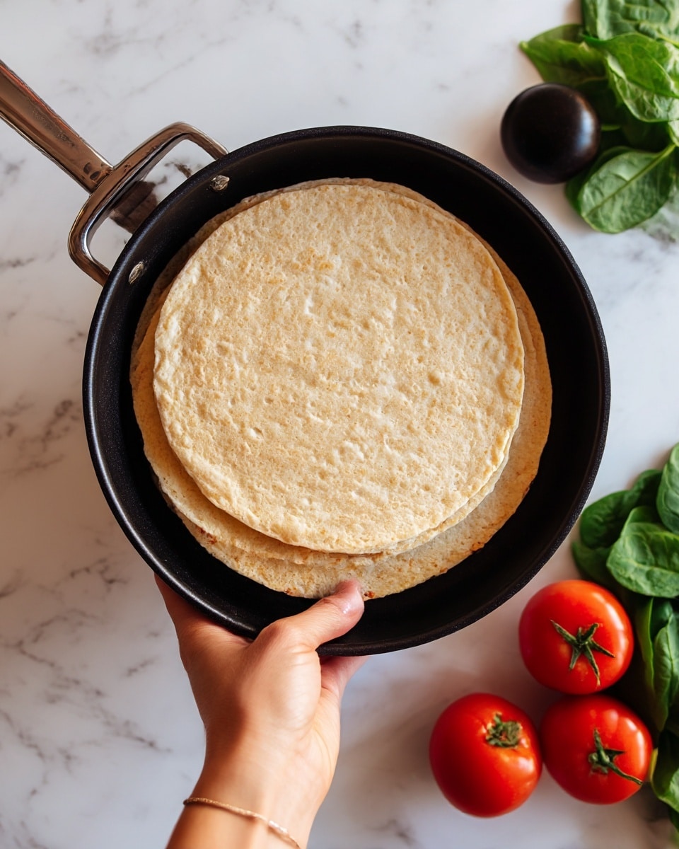 The image shows two round pizza crusts stacked on top of each other inside a black pan, resting on a white marbled surface. The crusts have a slightly golden and textured appearance, with a few air bubbles and uneven edges. Around the pan, there are fresh vegetables including three red tomatoes, one black tomato, and a bunch of green leafy spinach to the right. A woman’s hand is visible holding the pan handle on the left side. Photo taken with an iphone --ar 4:5 --v 7