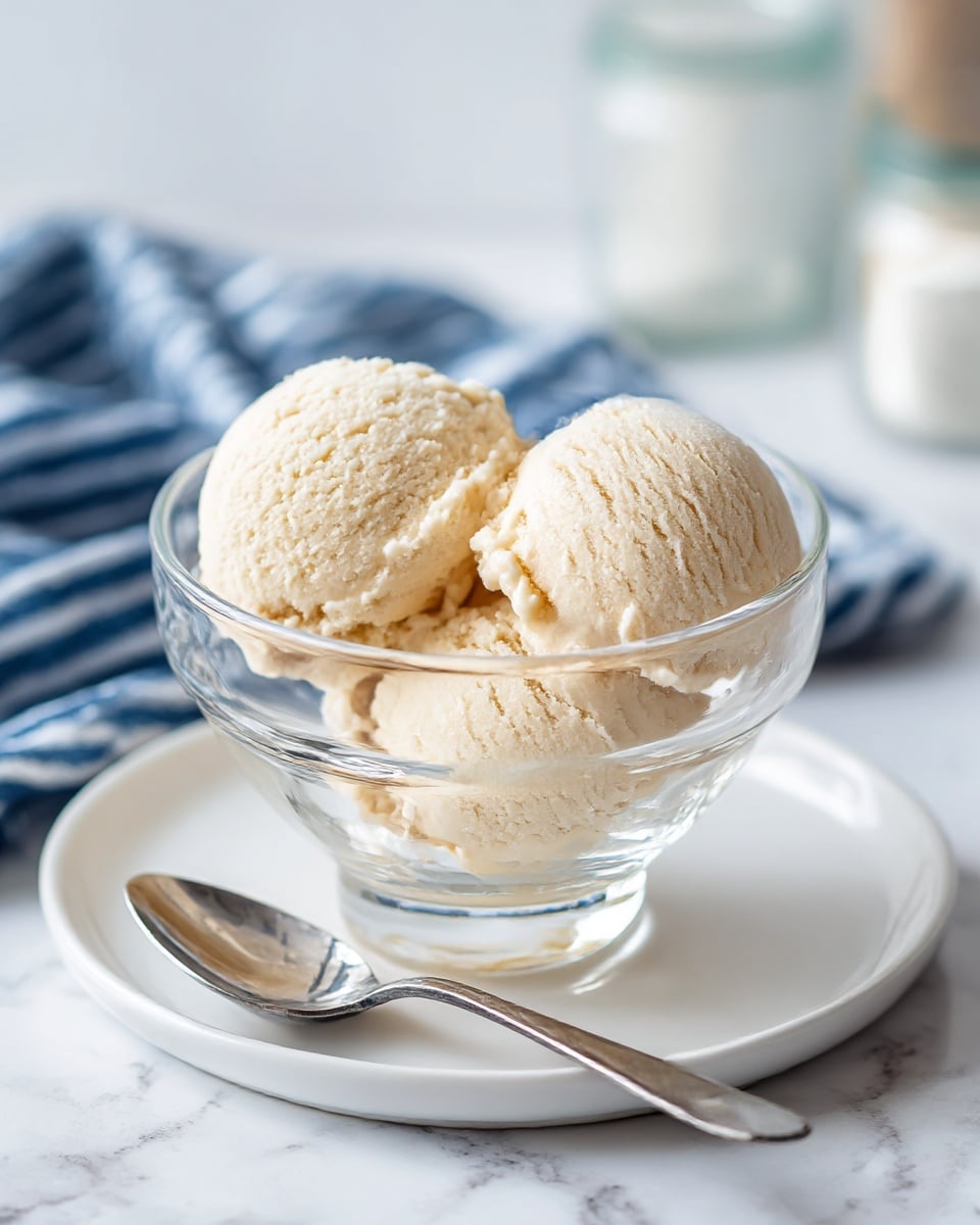 The image shows two scoops of light beige ice cream stacked inside a clear glass cup. The cup is placed on a shiny white plate with a silver spoon resting on the plate's edge to the right of the cup. The background features a white marbled texture, and behind the cup, there is a blurred checkered cloth in dark blue and white. The ice cream looks smooth and creamy with some texture visible on the scoops. photo taken with an iphone --ar 4:5 --v 7