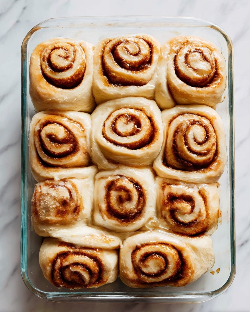 A clear glass baking dish holding six cinnamon rolls, with one roll missing, showing a soft, light tan dough spiraled with dark brown cinnamon filling, all covered with a thick, creamy white icing that looks smooth and slightly melted. A white spoon with a floral design rests inside the dish, coated in some icing and cinnamon. Next to the dish, on a white plate with a silver fork, there is a separated piece of a cinnamon roll showing soft layered dough and cinnamon in the middle, with some icing smeared on the plate. The setting is on a white marbled surface with a greenish dish towel partially visible. Photo taken with an iphone --ar 4:5 --v 7
