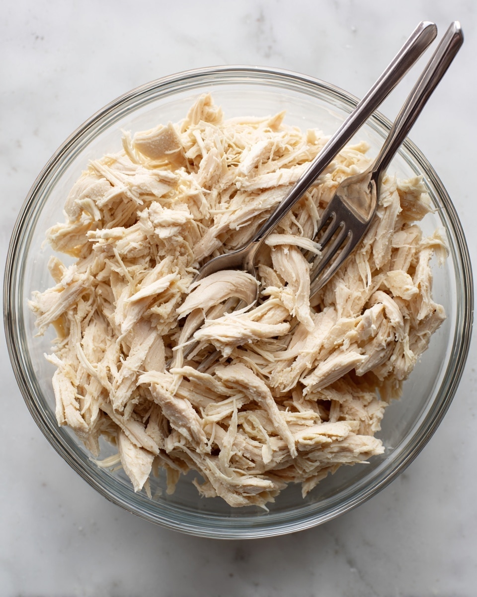 A clear glass bowl filled with shredded light beige cooked chicken pieces, showing soft and fibrous texture. Two silver forks are placed inside the bowl, one angled vertically near the top and the other diagonally on the right side, appearing to pull apart the chicken. The bowl sits on a white marbled surface that adds a clean and simple background with slight gray veining. photo taken with an iphone --ar 4:5 --v 7