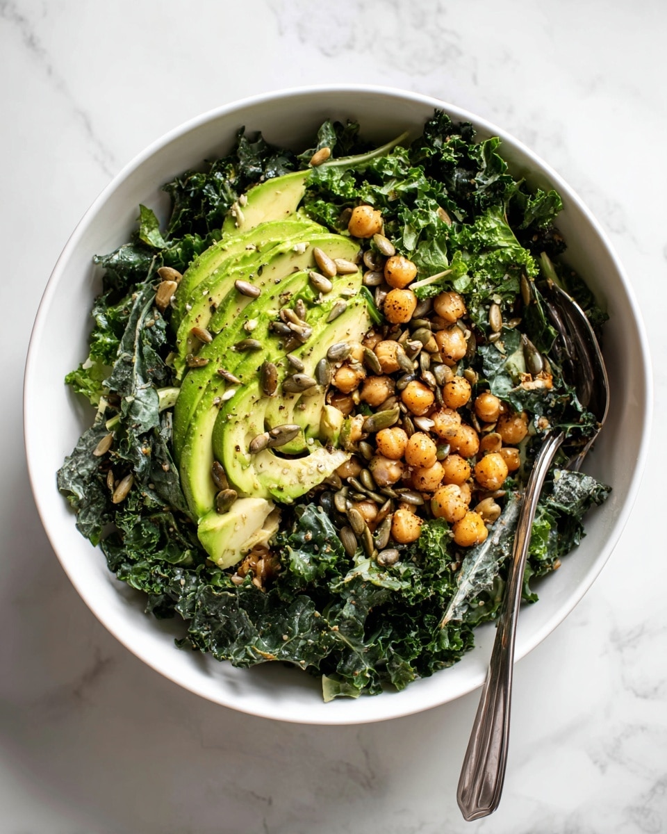 A white bowl filled with a fresh salad that has three main layers: the bottom layer is dark green kale leaves with a curly texture; the middle layer shows light beige chickpeas scattered evenly; the top layer has bright green, chunky slices of avocado mixed with small toasted brown sunflower seeds sprinkled lightly over the salad. A silver fork rests on the right edge of the bowl, partly inside the salad. The bowl sits on a white marbled surface. photo taken with an iphone --ar 4:5 --v 7