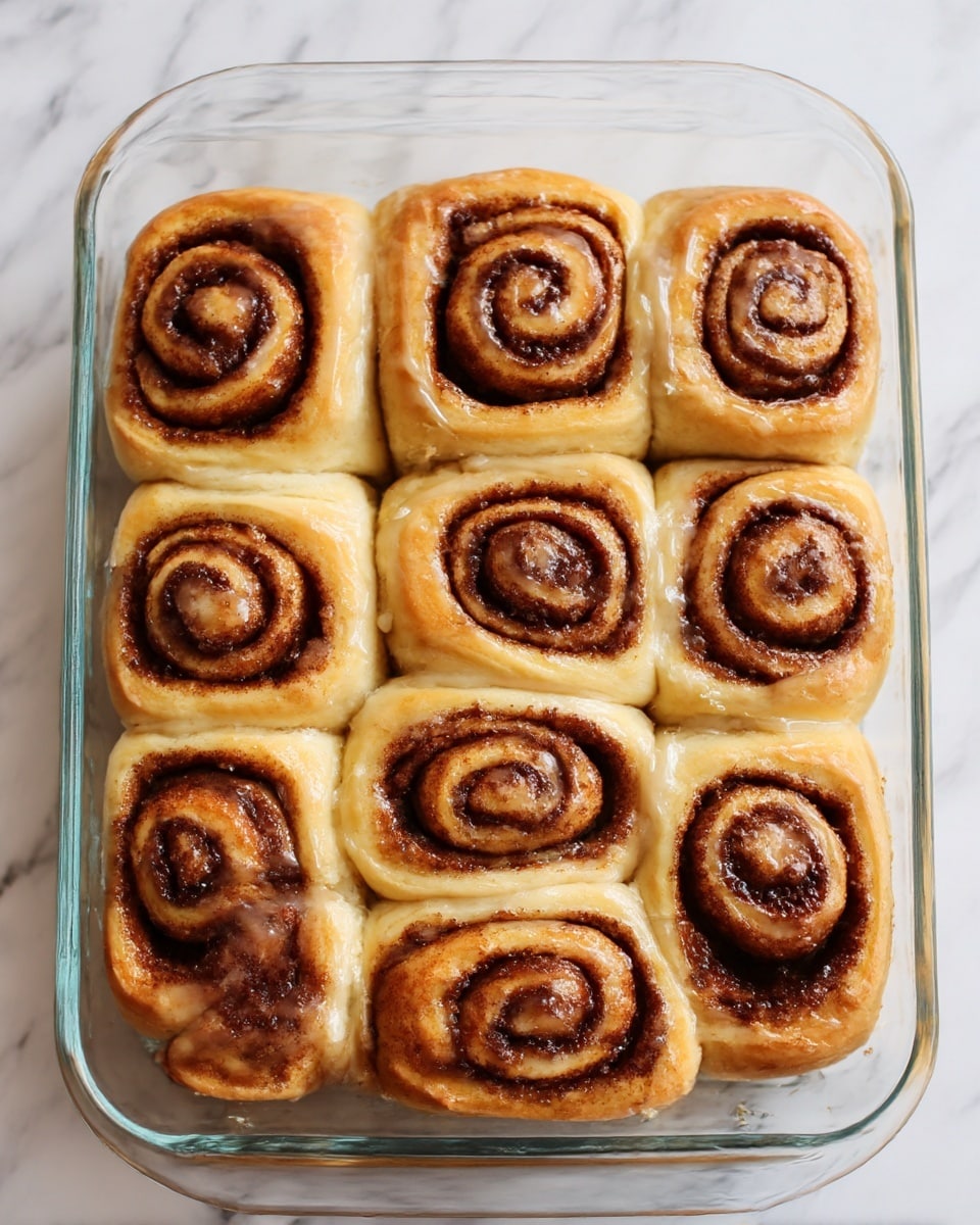 A glass baking dish filled with twelve cinnamon rolls arranged in a 4 by 3 grid, each roll showing a clear swirl pattern of brown cinnamon filling wrapped in soft, light beige dough with a slightly rough texture. The rolls are close together, touching each other, with some cinnamon filling slightly spilling between them, giving a sticky look on the sides. The background is a white marbled texture, and the photo is taken at a close angle, highlighting the spirals and the soft, fluffy dough. photo taken with an iphone --ar 4:5 --v 7