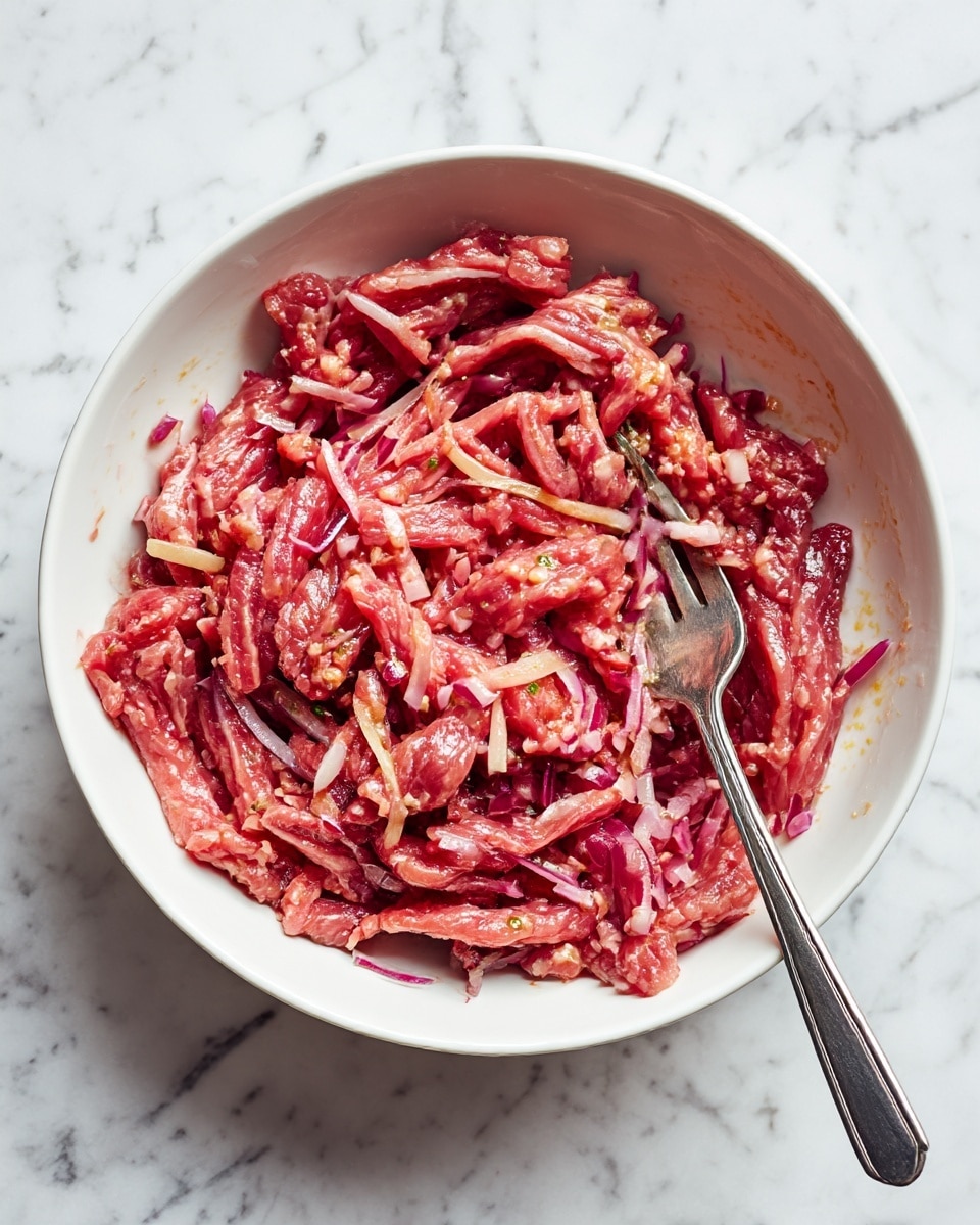 The image shows thin strips of raw meat with a pinkish-red color, mixed with small pieces of finely chopped red onions. The mixture sits inside a white bowl with a silver fork placed in it, visible on the right side. The texture of the meat looks soft and moist, while the onions add a slightly translucent and shiny layer on top. The white bowl rests on a white marbled surface, creating a clean and bright visual setting. Photo taken with an iphone --ar 4:5 --v 7