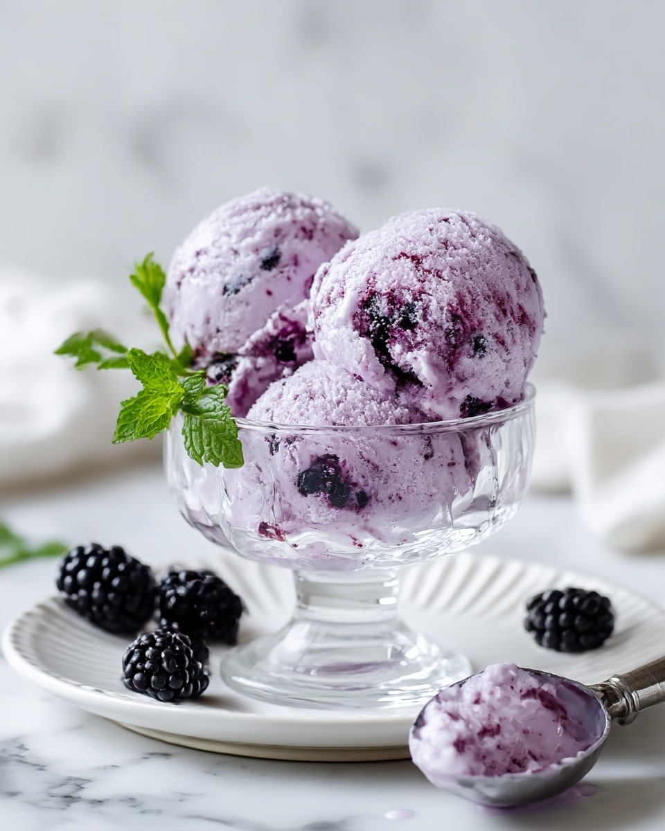 Two scoops of light purple ice cream with dark purple berry pieces mixed inside are placed in a clear glass dessert cup, which sits on a white plate with a ridged edge on a white marbled surface. There is a small sprig of fresh green mint on the left side of the ice cream. In the background, two blackberries lay on the white plate near the base of the glass, and a silver ice cream scoop with some melted purple ice cream is sitting on the surface in the foreground. Photo taken with an iphone --ar 4:5 --v 7