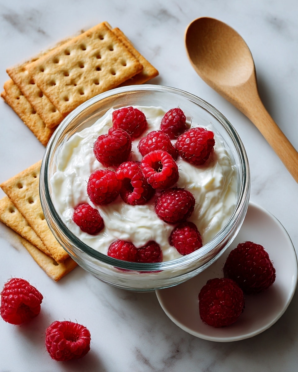 A clear glass bowl sits on a white marbled surface filled with a creamy white mixture topped with bright red raspberries, giving a fresh and smooth texture contrast. To the top right of the bowl is a white plate with two raspberries laying on it, showing a close view of the berry's textured surface. On the top left near the bowl, two rectangular golden brown crackers rest flat on the white marbled surface, their small holes and slight sheen visible. A wooden spoon with a smooth pale brown texture lies vertically to the right side, adding a natural touch to the simple kitchen scene. photo taken with an iphone --ar 4:5 --v 7