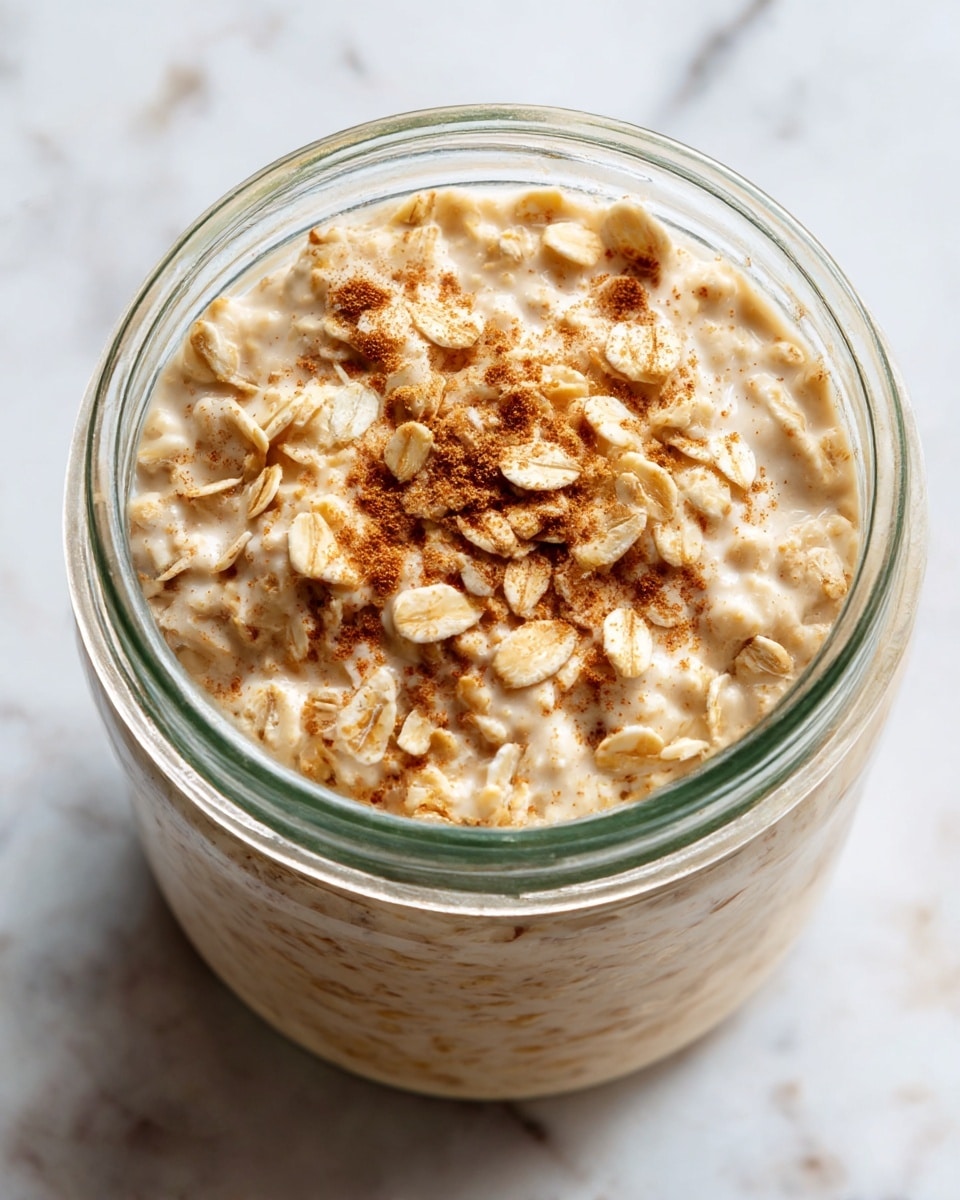 A clear glass jar filled with thick, creamy oatmeal that has a light beige color with visible oats mixed throughout. The surface is sprinkled lightly with small bits of cinnamon powder giving a soft brown contrast on the oatmeal. The jar sits directly on a white marbled surface with natural light highlighting the smooth and textured parts of the oats, showing both the soft and still slightly whole rolled oats on top. Photo taken with an iphone --ar 4:5 --v 7