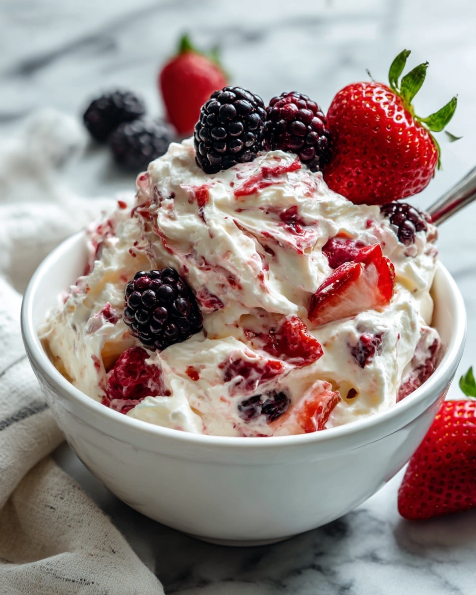A white bowl filled with a creamy mixture that has white whipped cream swirled thickly with pieces of red strawberries, black blackberries, and red raspberries mixed in. The cream and berries create a fluffy, textured surface with the bright colors of the fruit showing through the white. On top, a few whole berries—blackberry, raspberry, and strawberry—sit close together adding a pop of color to the creamy mix. The bowl is set on a white marbled surface with a folded cloth nearby, giving a fresh and simple look. photo taken with an iphone --ar 4:5 --v 7