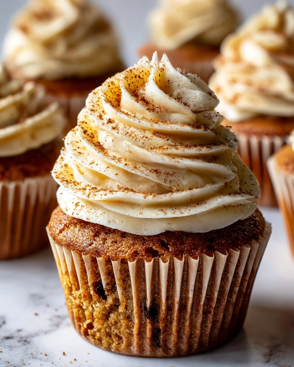 The image shows five cupcakes arranged on a round wooden board placed on a white marbled surface. Each cupcake has one layer of brown cake base with small dark spots and one layer of thick, creamy white frosting swirled on top, dusted lightly with a brown powder. In the background, there is a jar of amber-colored jam with a spoon inside, and to the right of the cupcakes, two figs are present—one whole purple fig and one fig cut in half showing red and pink inner flesh with seeds. The overall scene is bright with a soft light and neutral tone. photo taken with an iphone --ar 4:5 --v 7