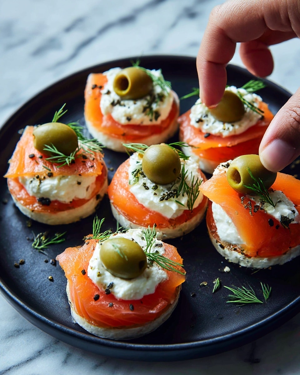 Six small rolls are placed on a black round plate, each made of a first layer of thin, orange smoked salmon wrapped tightly. On top of the salmon, there is a thick, white creamy dollop, smooth in texture, and centered with a green olive slice. Small sprigs of fresh green dill are arranged around the olive slice like a star. A woman's hand is seen picking up one of the rolls from the bottom left of the plate. The plate rests on a white marbled surface. photo taken with an iphone --ar 4:5 --v 7